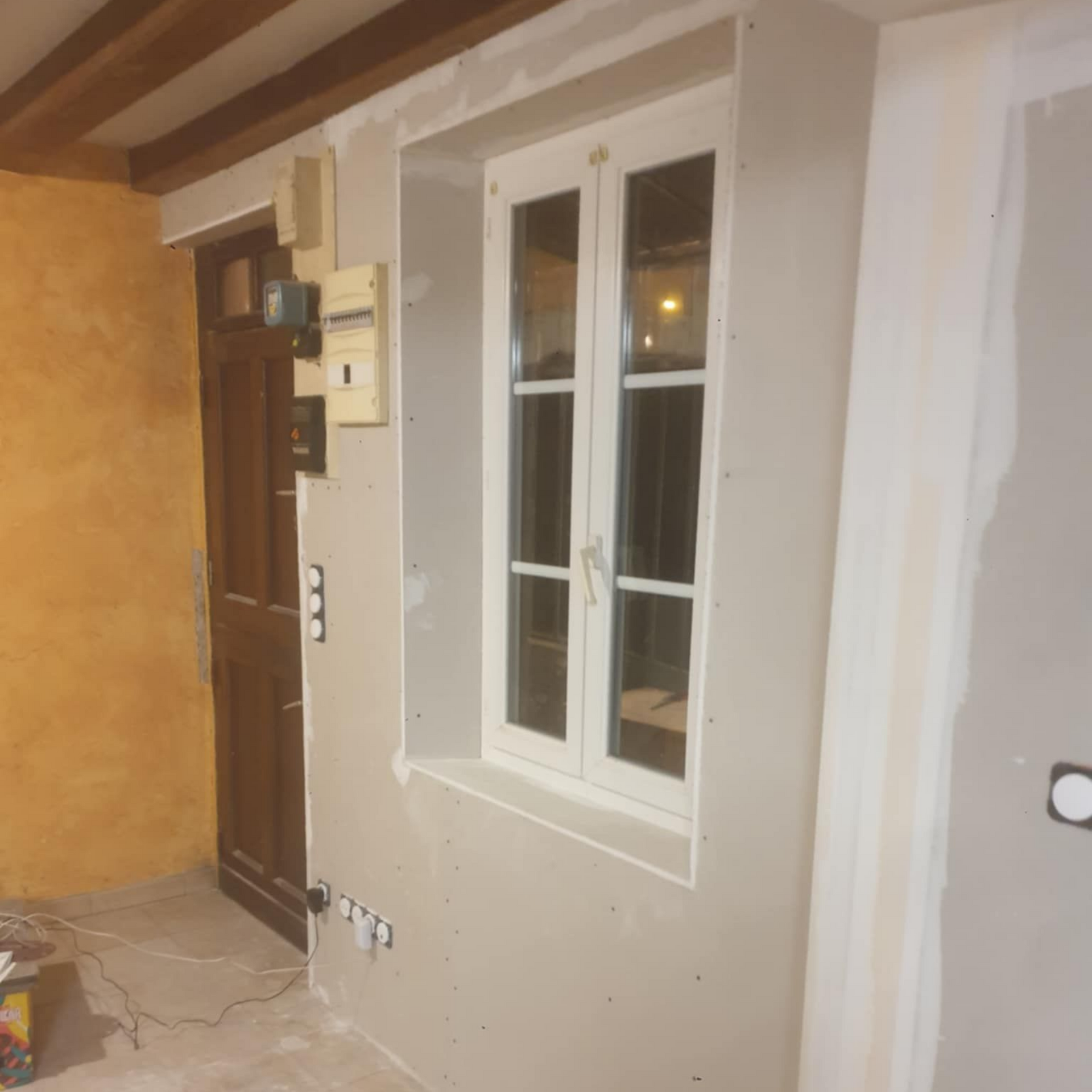 Rénovation en cours : une fenêtre, une porte et un panneau électrique sur un mur de couleur claire ; crépi brut et poutres apparentes.