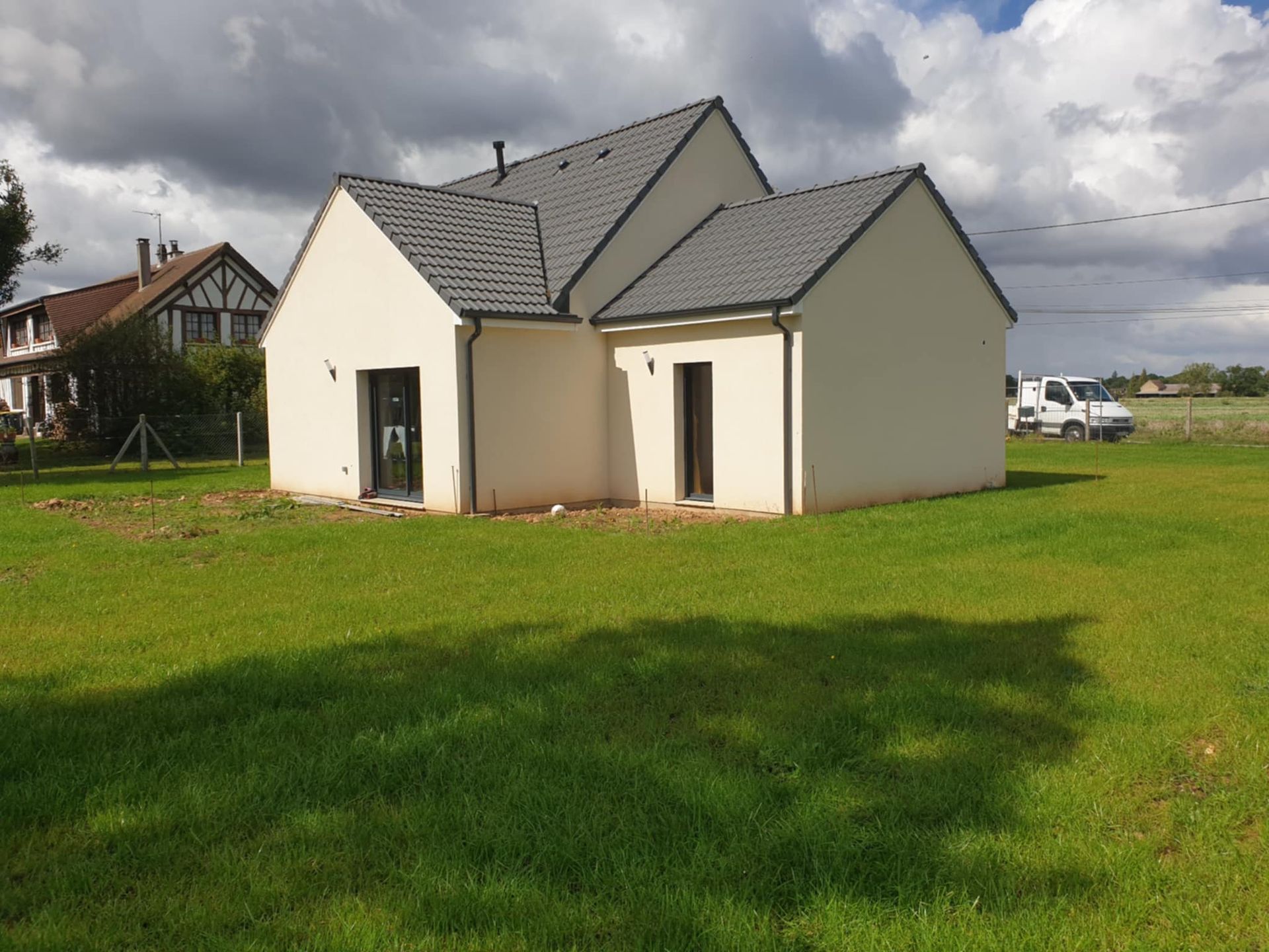 Maison beige moderne avec toit gris foncé, sur herbe verte, ciel nuageux.