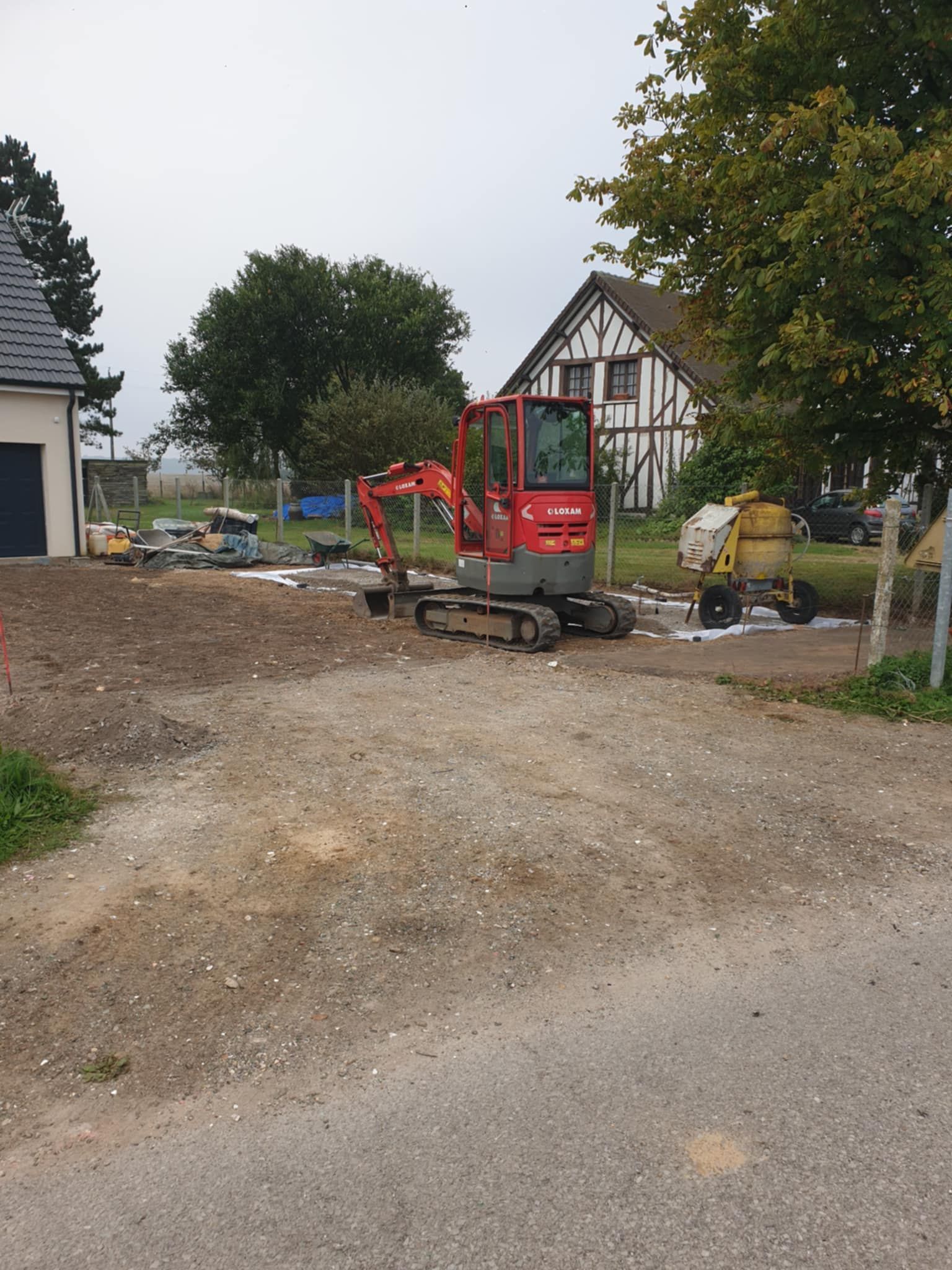 Excavatrice rouge sur un chemin de gravier ; chantier de construction avec des maisons en arrière-plan.