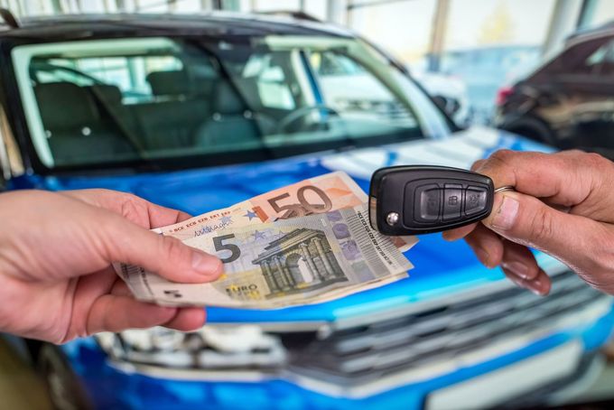Una persona sostiene una llave de coche y un fajo de dinero delante de un coche.