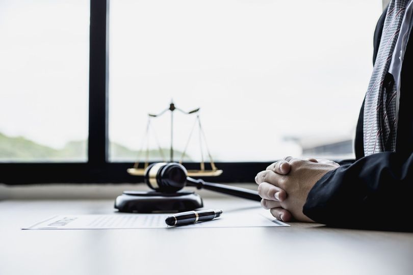 Las manos de una persona unidas delante del mazo de un juez y una balanza de la justicia sobre un escritorio.