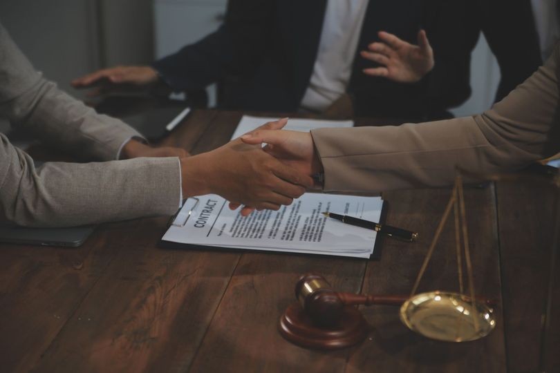 Personas se dan la mano para firmar un contrato en una mesa con una balanza de la justicia y un mazo.