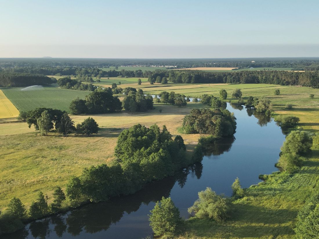 Der Fluss schlängelt sich unter klarem Himmel durch grüne Felder und Bäume.