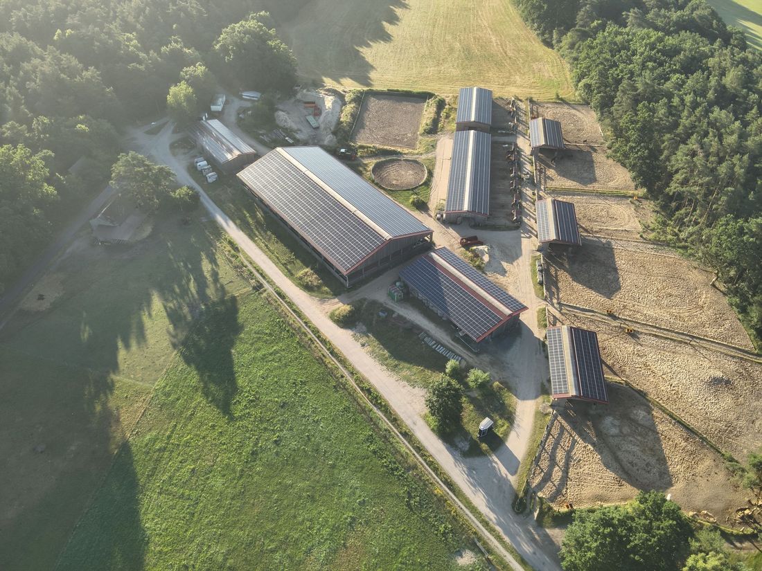 Luftaufnahme eines Bauernhofs mit Gebäuden, Solarmodulen auf den Dächern und umliegenden Feldern und Bäumen.
