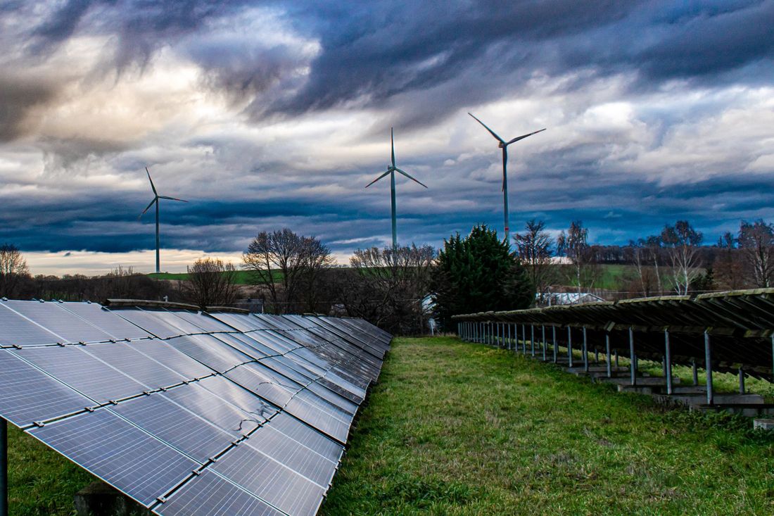 Solarmodule auf einer Wiese mit Windmühlen im Hintergrund, unter einem bewölkten Himmel.