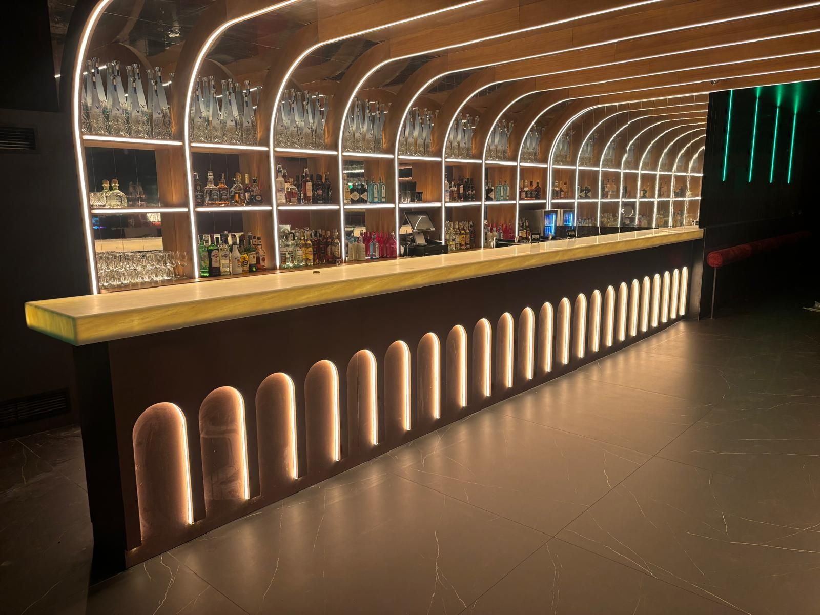 Interior de bar moderno con estantes arqueados iluminados, botellas y cálidos detalles de madera.