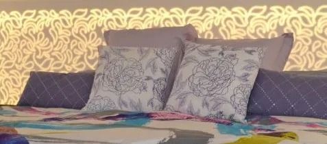 Cama con almohadas grises estampadas y una colorida manta floral sobre un cabecero floral amarillo.