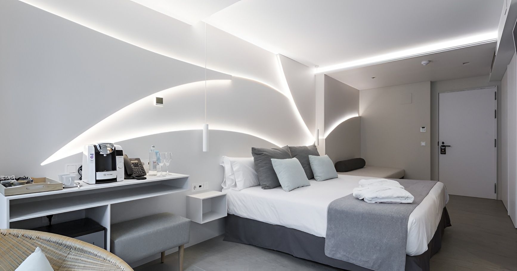 Habitación de hotel moderna de color blanco con dos camas individuales, escritorio empotrado, iluminación LED curva y puerta de baño en la parte trasera.