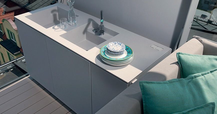 Mesa de patio exterior con cristalería transparente, una pila de platos y cojines color turquesa a su lado.