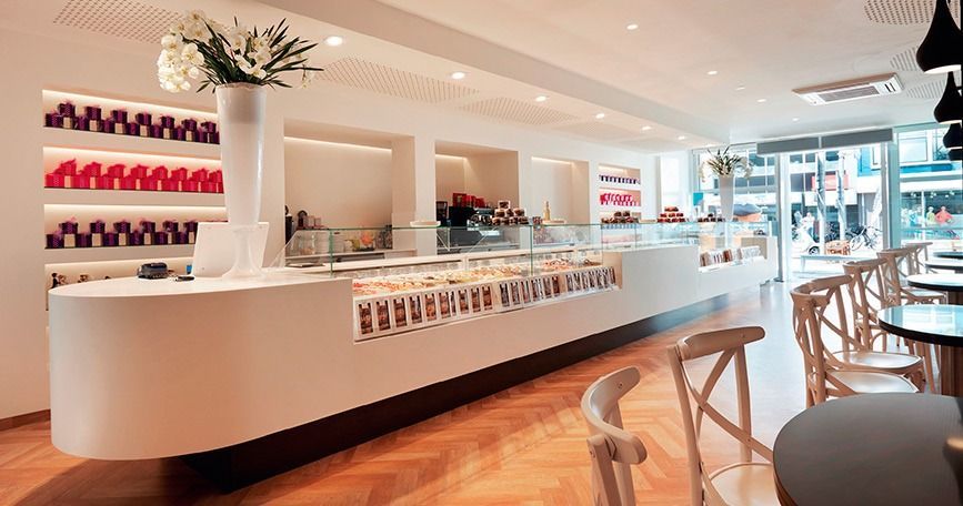 Interior de cafetería moderno y luminoso con mostradores blancos, sillas de madera y zona de estar soleada.
