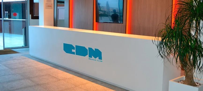 Mostrador de recepción de oficina moderno con logotipo azul, ventanas de cristal y una planta en maceta con iluminación cálida.