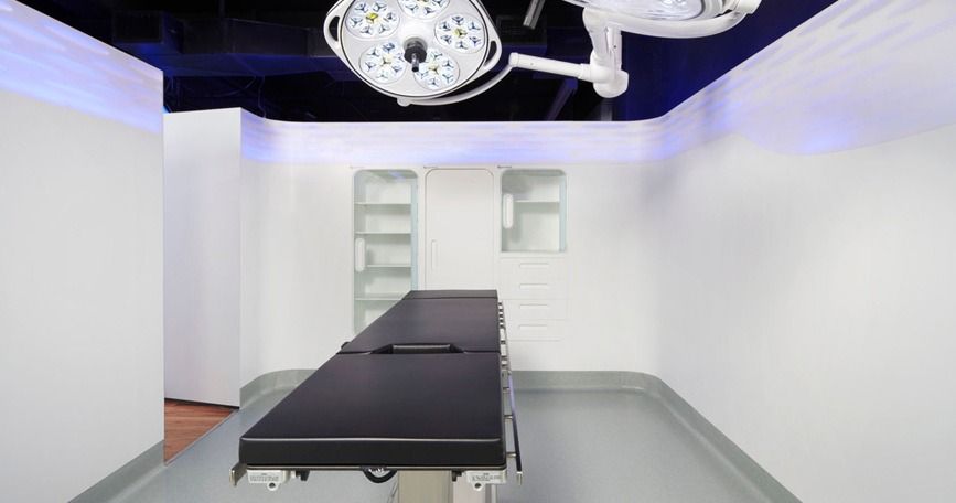 Consultorio médico moderno con camilla negra, paredes blancas, luces de techo y detalles de iluminación púrpura.