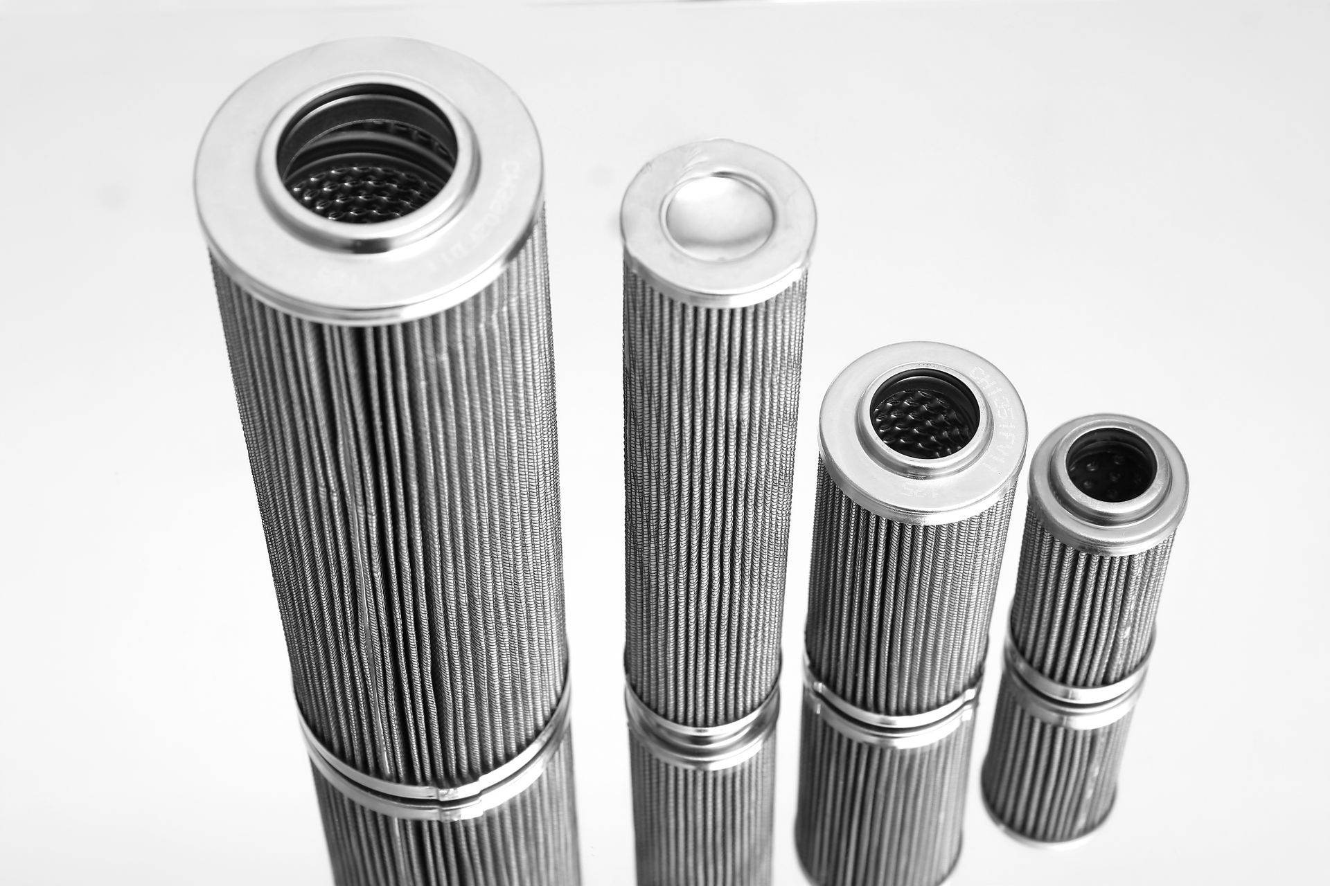 Quatre filtres métalliques industriels cylindriques de tailles différentes sont disposés verticalement sur une surface réfléchissante, sur fond blanc.