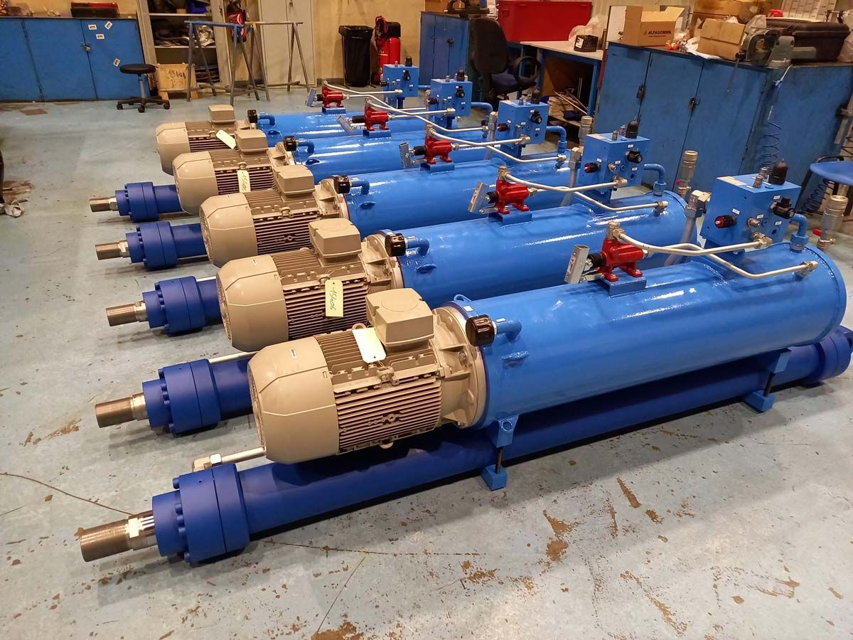 Une rangée de vérins hydrauliques industriels bleus identiques, équipés de moteurs beiges, sont disposés sur le sol en béton d'un atelier.