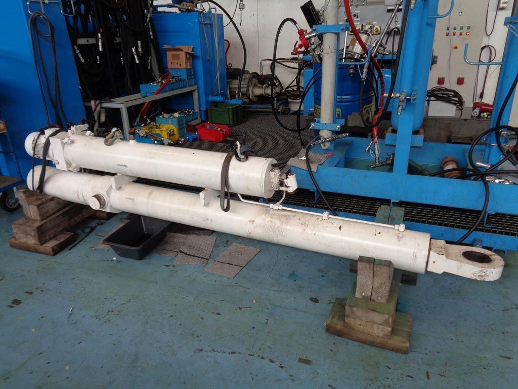 Deux gros vérins hydrauliques industriels blancs reposent sur des blocs de bois dans un atelier.