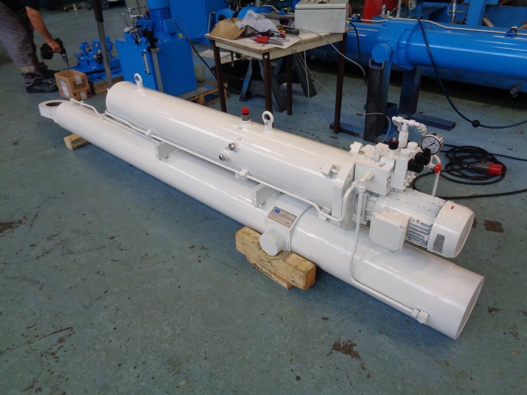 Un gros vérin hydraulique industriel blanc, avec un moteur et un ensemble de vannes intégrés, reposant sur des blocs de bois dans un atelier.