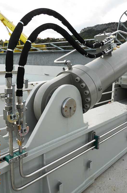 Un vérin hydraulique, peint en gris clair, avec des tuyaux noirs et des tubes en acier, monté sur une structure.