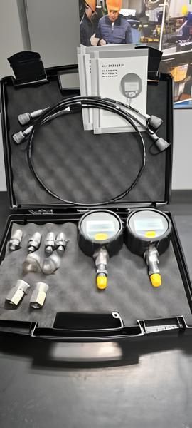 Une mallette noire ouverte contenant deux manomètres numériques, des tuyaux, des manuels et des raccords.