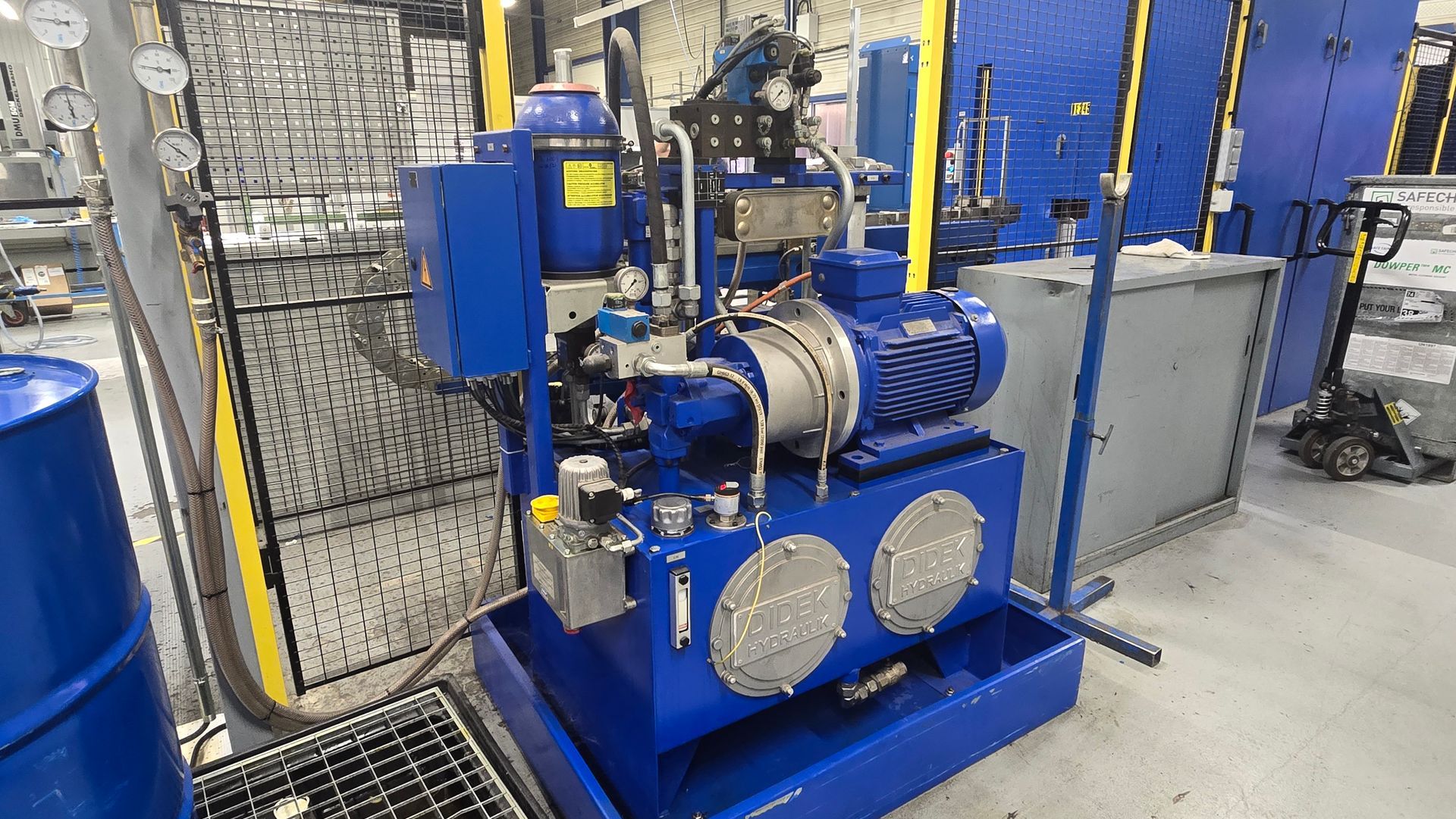 Une centrale hydraulique industrielle bleue est posée sur le sol d'une usine, à côté d'un fût bleu et de machines.