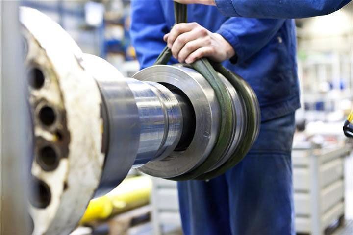 Un ouvrier en salopette bleue manipule une grosse pièce métallique à l'aide d'une sangle de levage dans une usine.