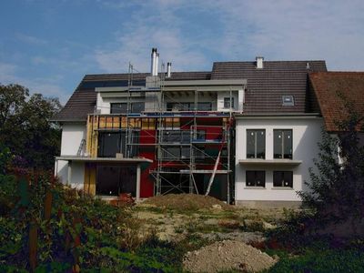 Ein im Bau befindliches Haus mit Gerüst davor, weisser Fassade und dunklem Ziegeldach vor klarem, blauem Himmel.