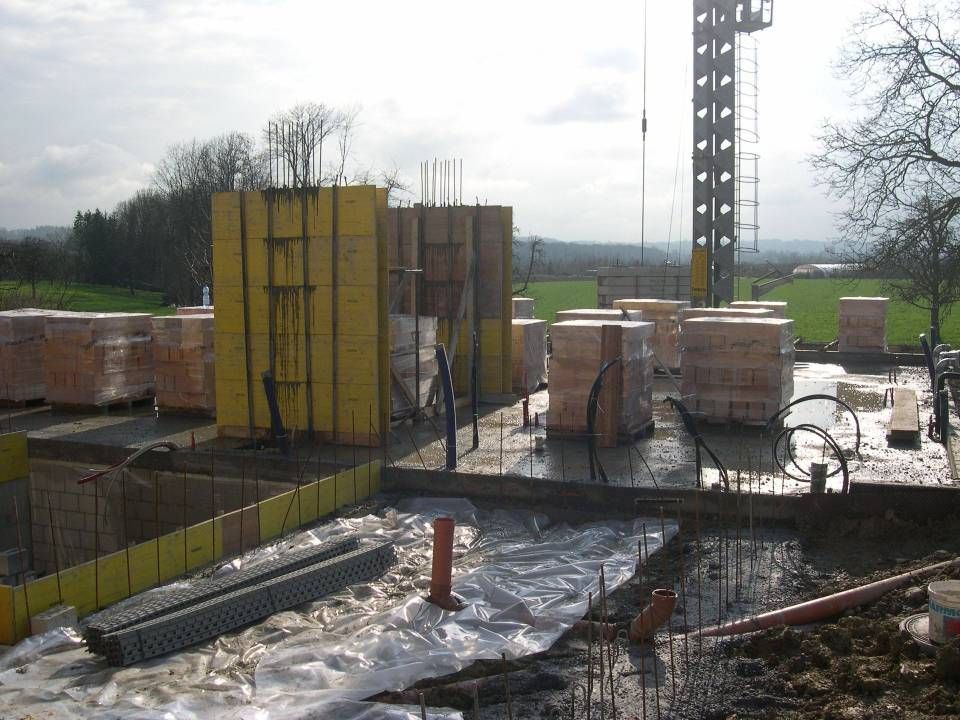 Eine Baustelle mit Betonfundamenten, gelber Schalung, gestapelten Paletten mit Ziegelsteinen und einem Turmdrehkran.