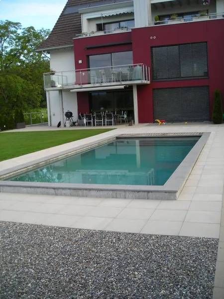 Auf einer Terrasse neben einem modernen Haus mit roten Wänden und einem Balkon befindet sich ein rechteckiger Swimmingpool.