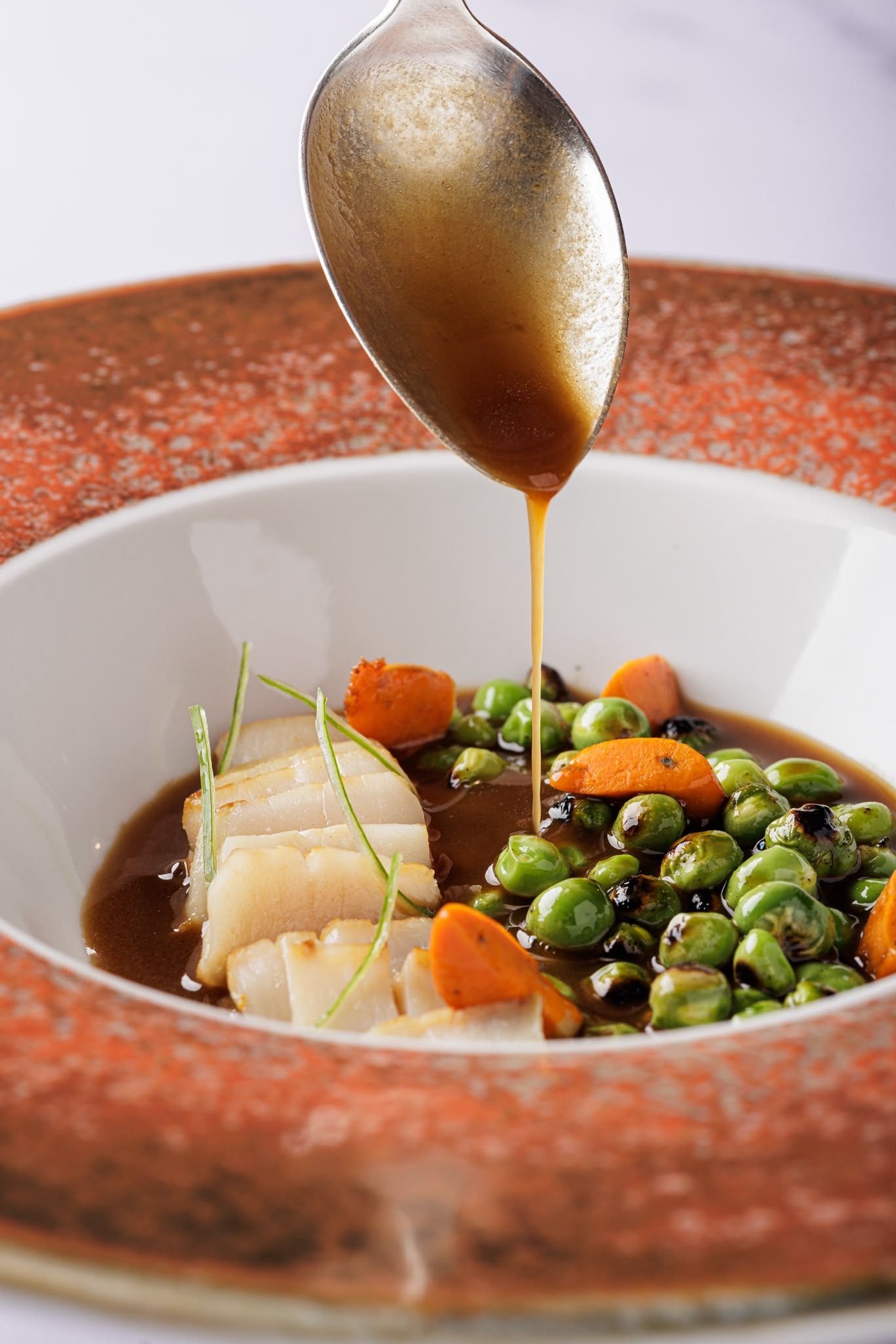 Sopa servida en un cuenco blanco con guisantes y verduras, mientras se vierte salsa con una cuchara sobre un plato rojo.