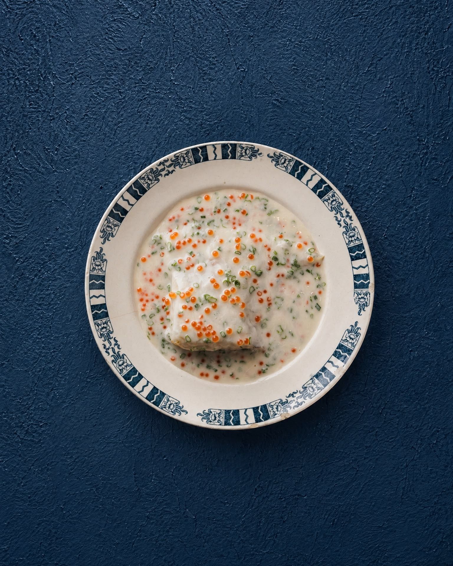 Un cuenco de sopa cremosa con hierbas y especias sobre un plato estampado, con un fondo azul oscuro
