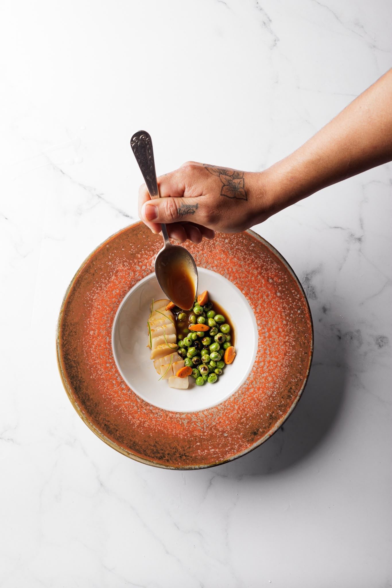 Una mano colocando salsa sobre un plato servido en un cuenco con borde de cobre, sobre una superficie blanca