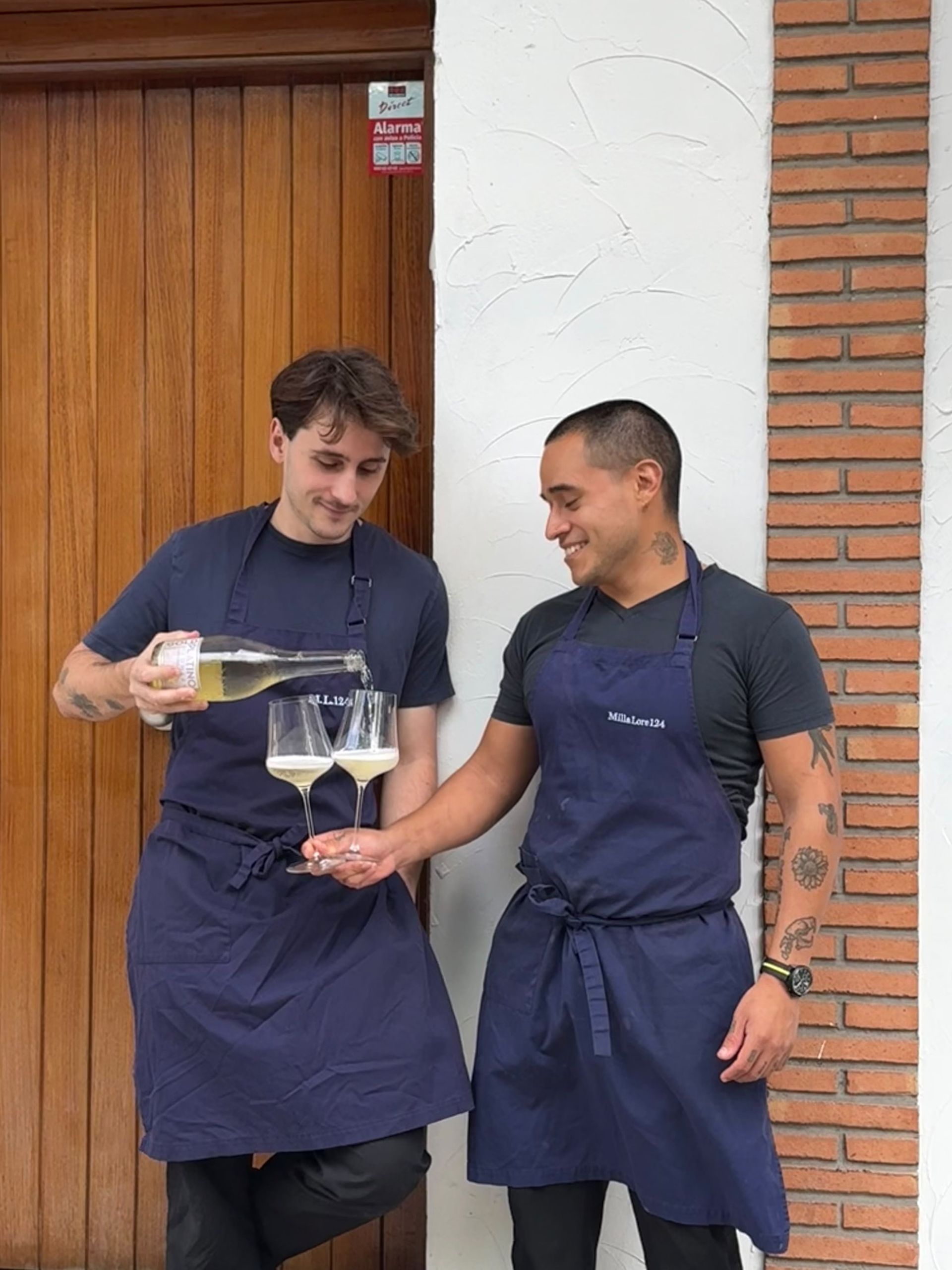 Dos camareros con delantales oscuros sirven vino juntos junto a una puerta de madera.