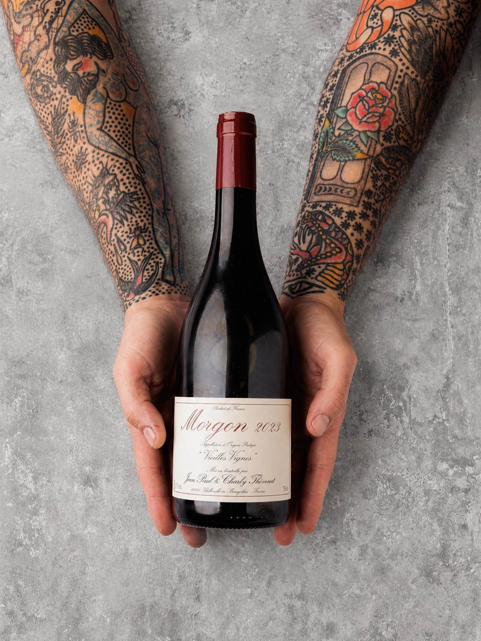Brazos tatuados sosteniendo una botella de vino tinto oscuro con una etiqueta blanca sobre un fondo gris