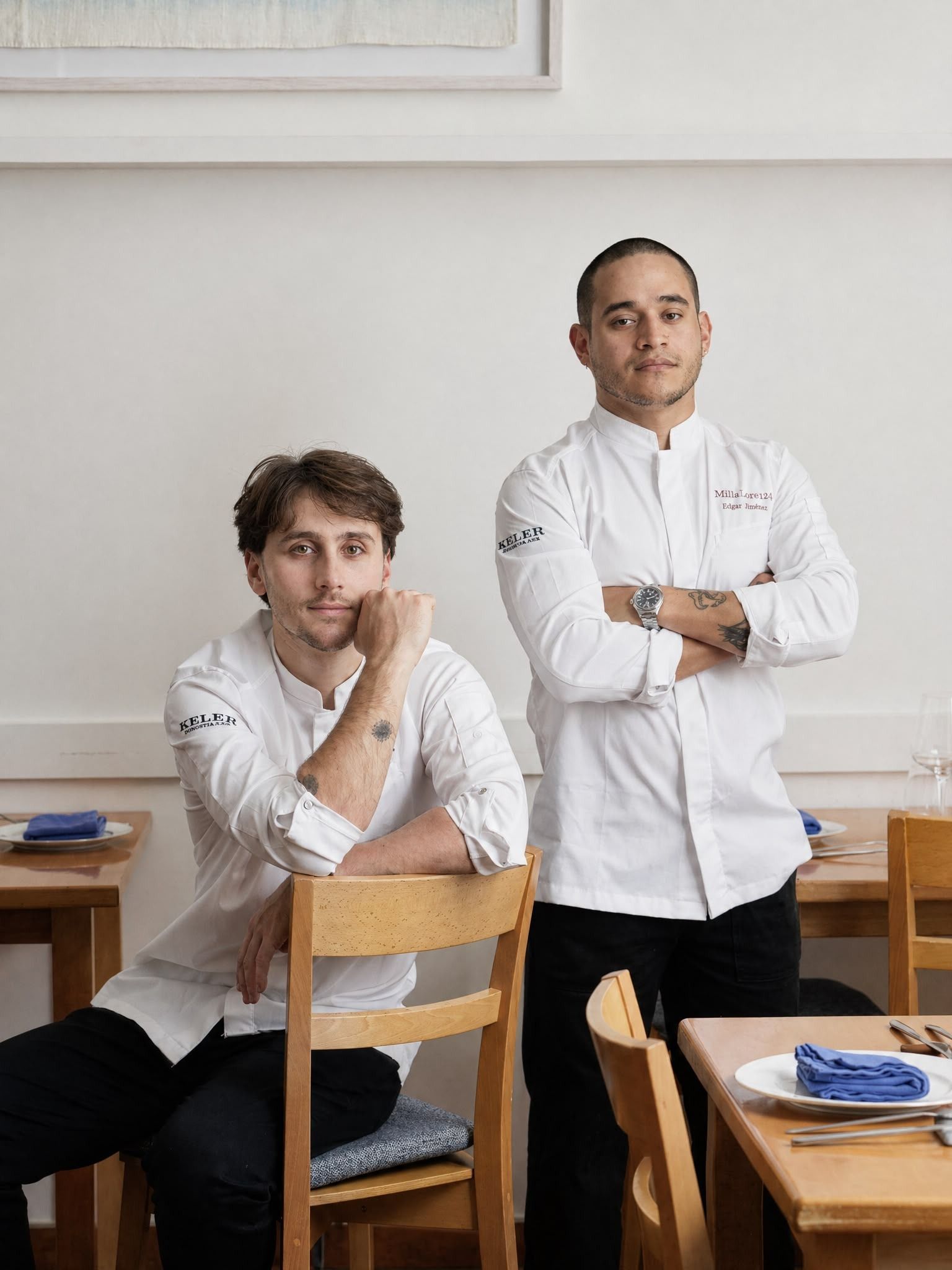 Dos personas con chaquetas blancas de chef posan en un comedor con sillas de madera y servilletas azules sobre las mesas.