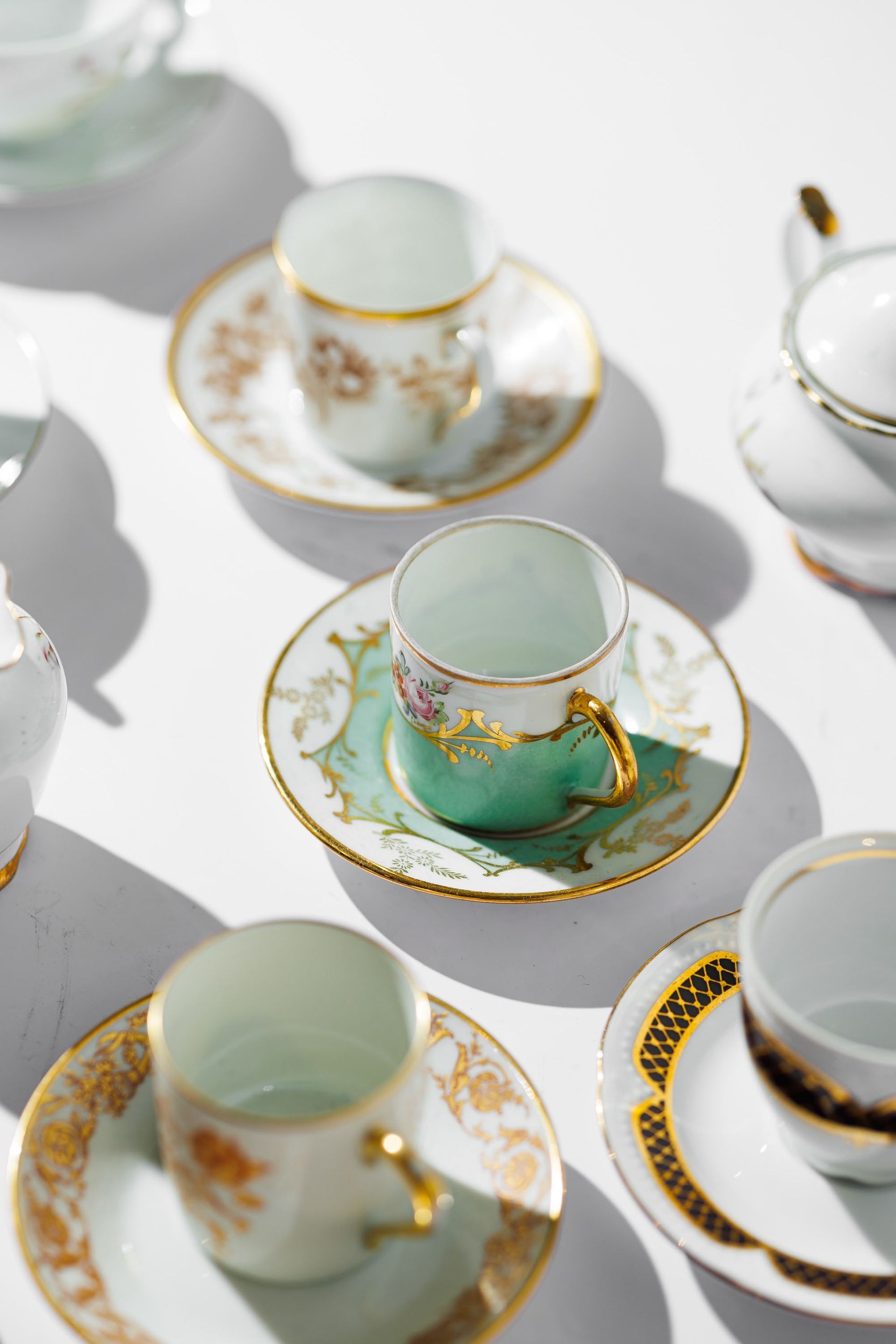 Tazas de té y platillos variados con ribetes dorados, sobre una superficie blanca y con motivos en verde menta y blanco