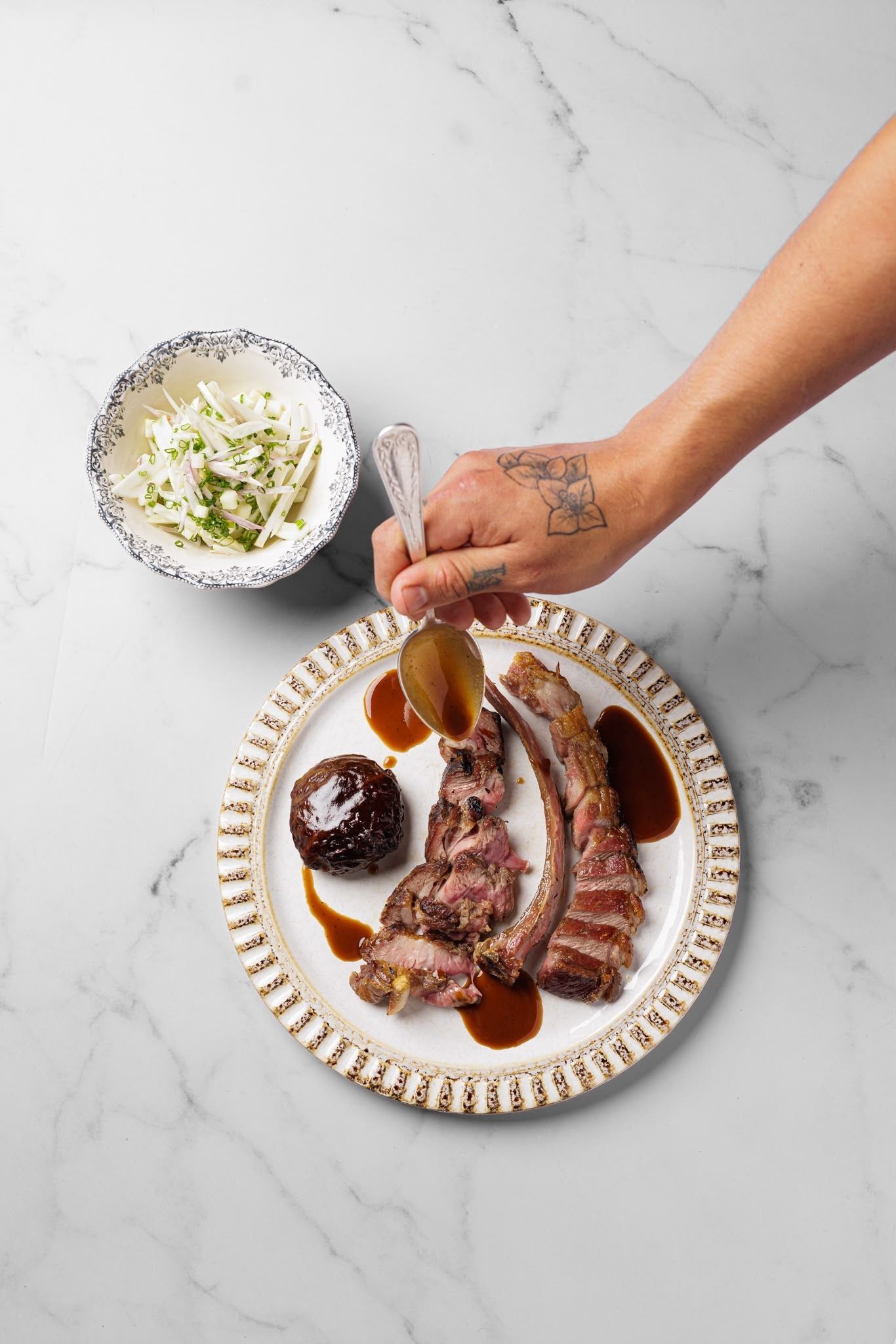 Emplatando a mano filete en rodajas con salsa, junto a una ensalada sobre una mesa de mármol blanco