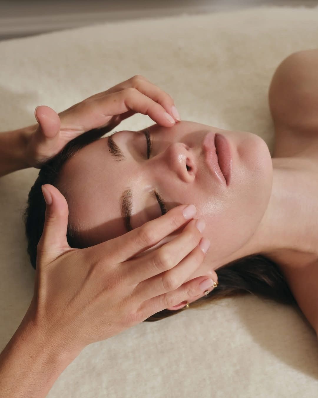 Des mains massent le visage d’une personne allongée sur une table de spa, les yeux fermés.