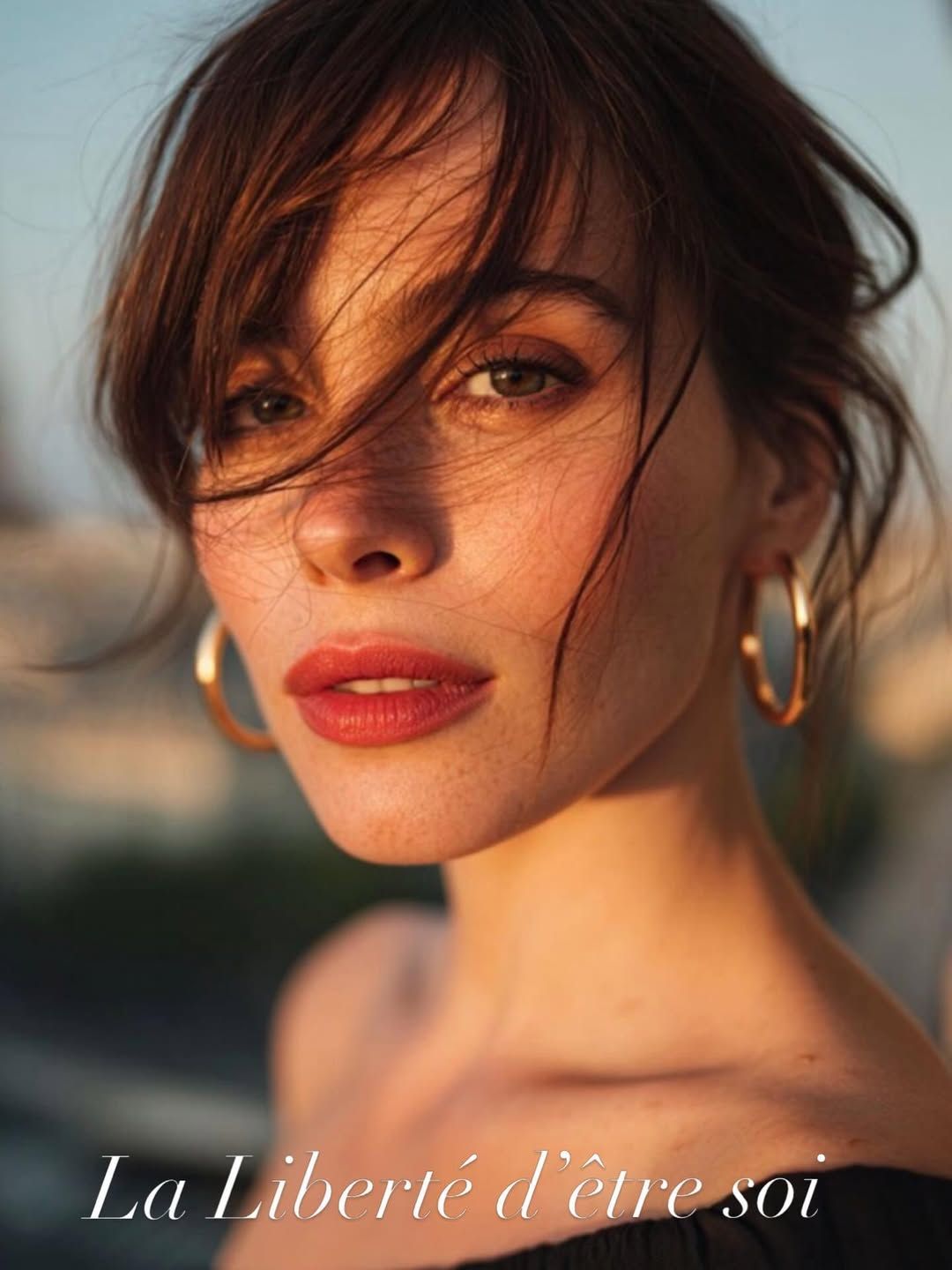 Portrait en gros plan d’une femme aux cheveux ébouriffés par le vent et portant des créoles au coucher du soleil, avec le texte « La liberté d’être soi »