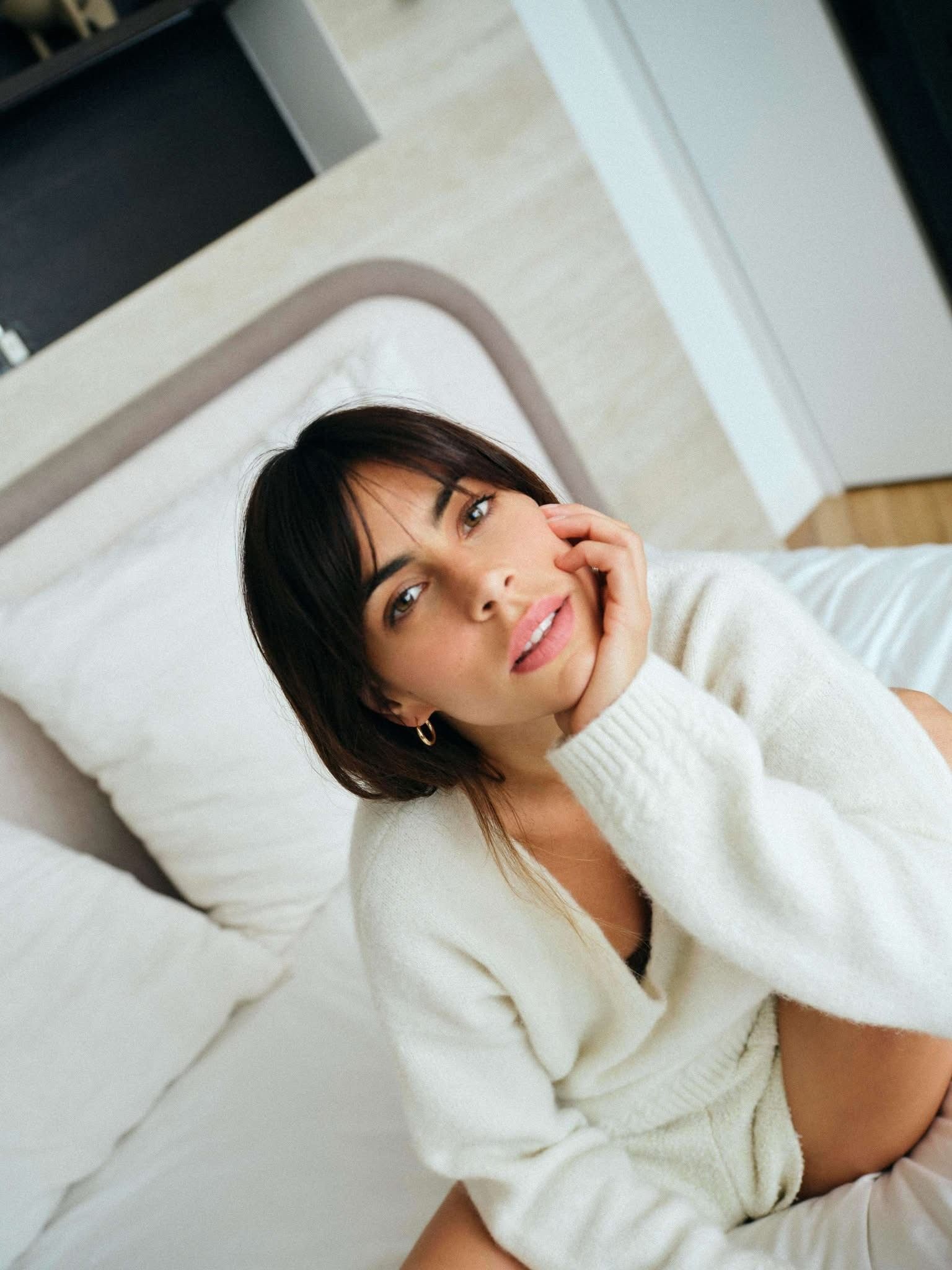 Femme en pull blanc allongée sur un lit, regardant l’appareil photo, la main sur le visage.