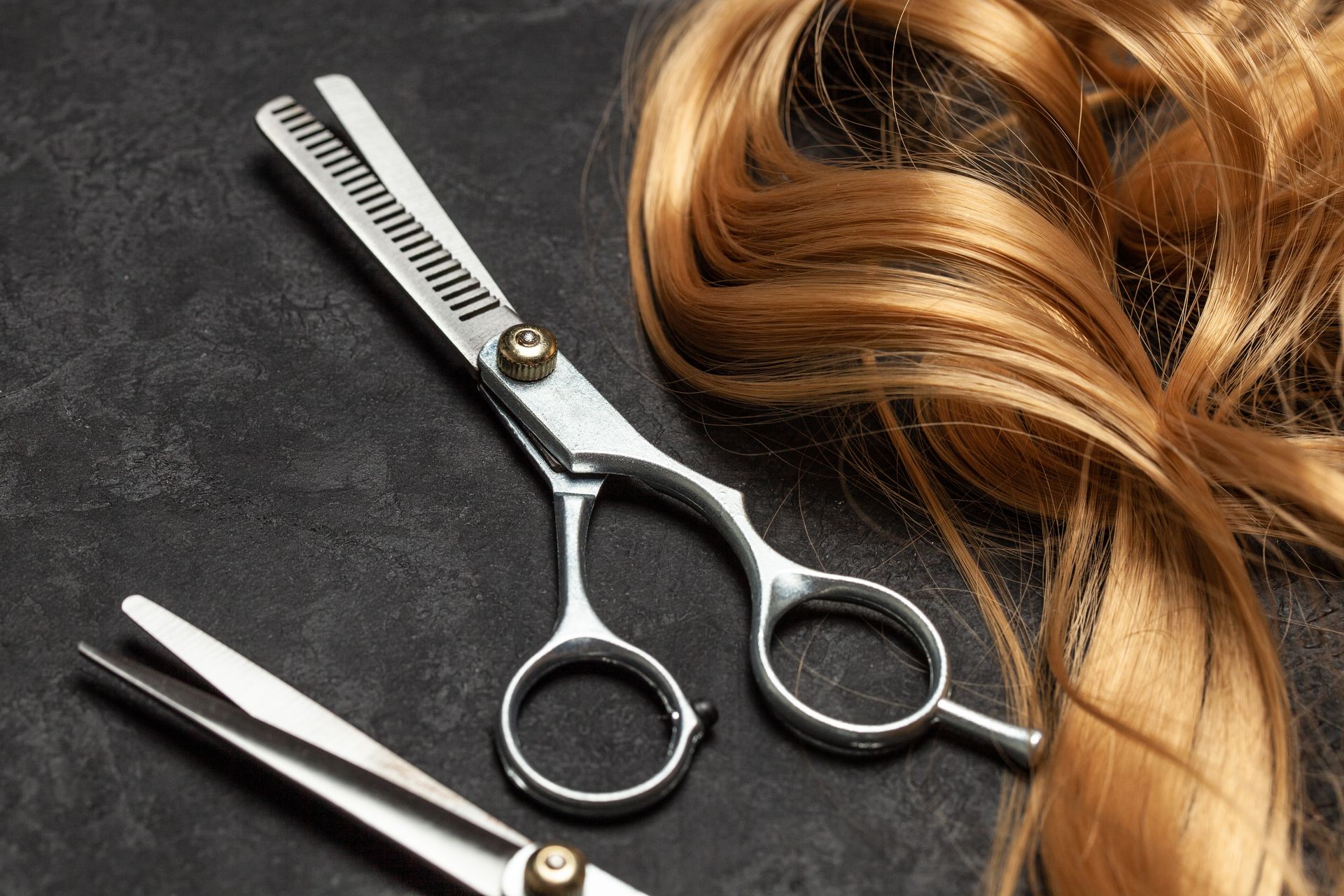 Scheren und lockiges, blondes Haar auf einem grauen Hintergrund: Wir schneiden und stylen Ihren neuen Haarersatz.