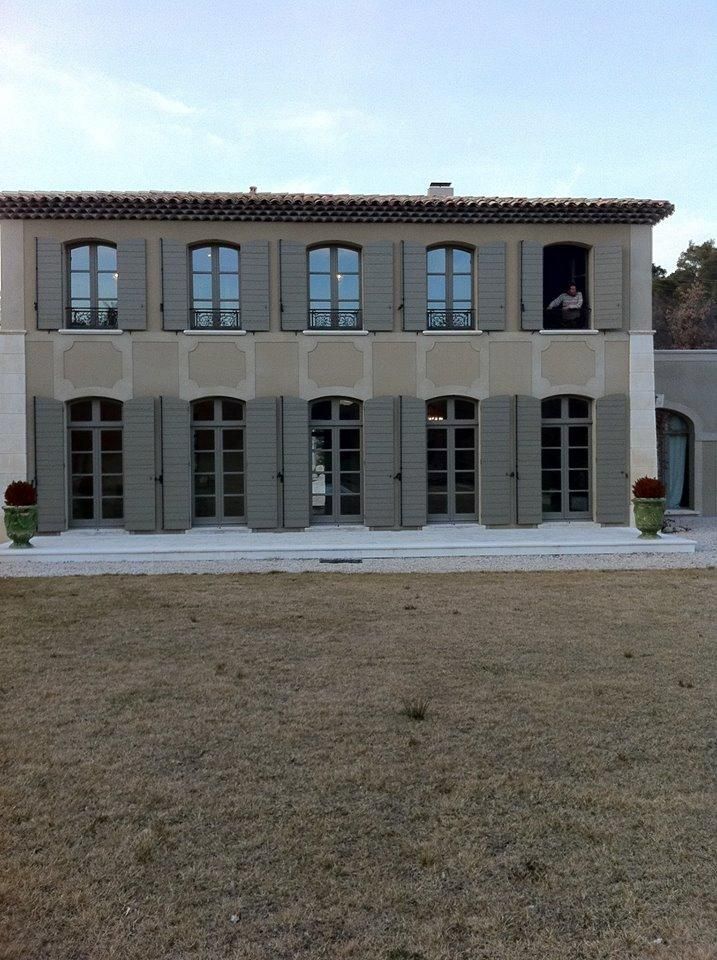 Maison beige à deux étages avec volets et fenêtres gris ; personne à la fenêtre, cour avant avec herbe sèche.