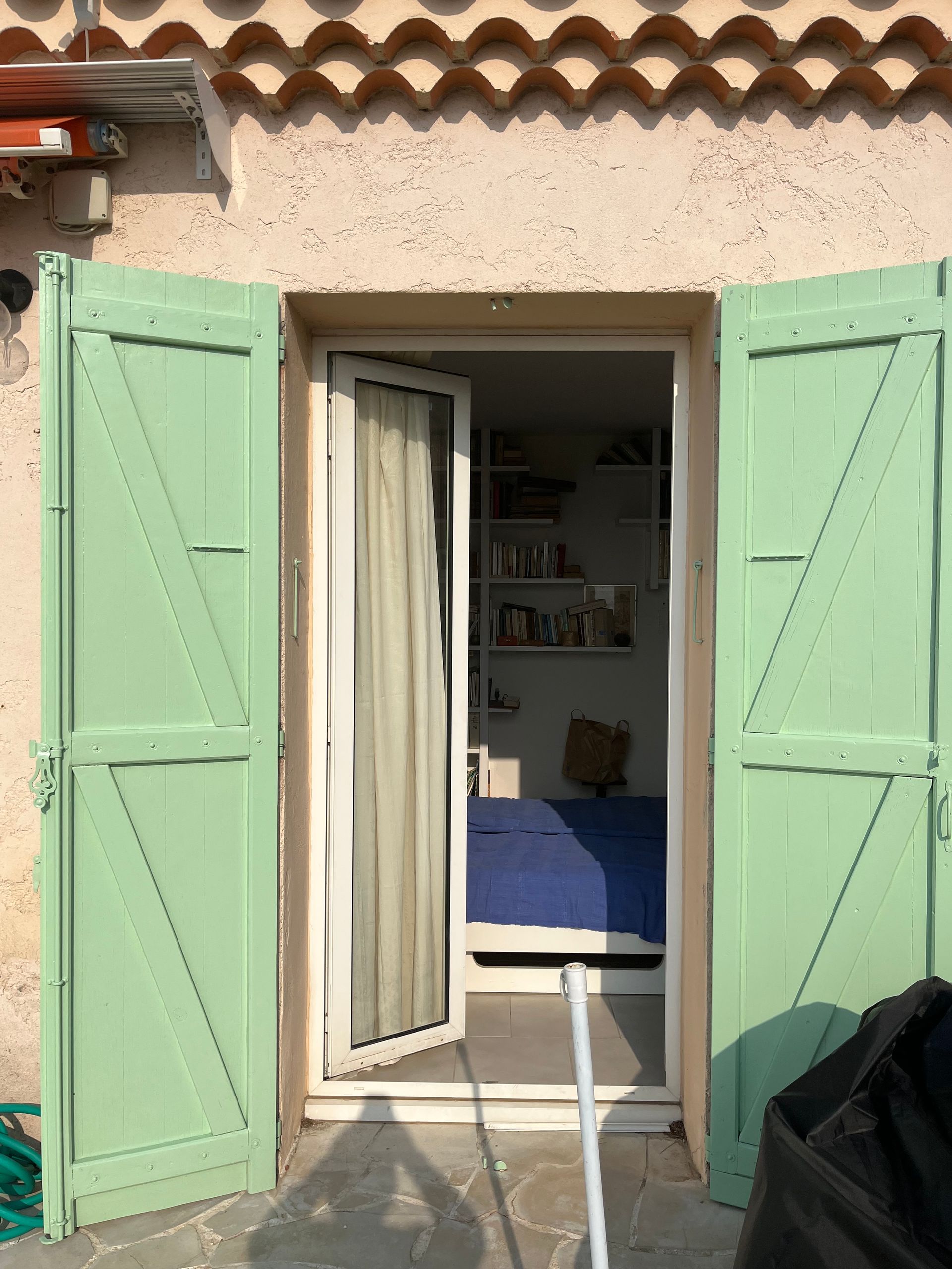 Des volets vert clair encadrent une porte ouverte. Cette porte donne sur une chambre avec un lit et une bibliothèque.