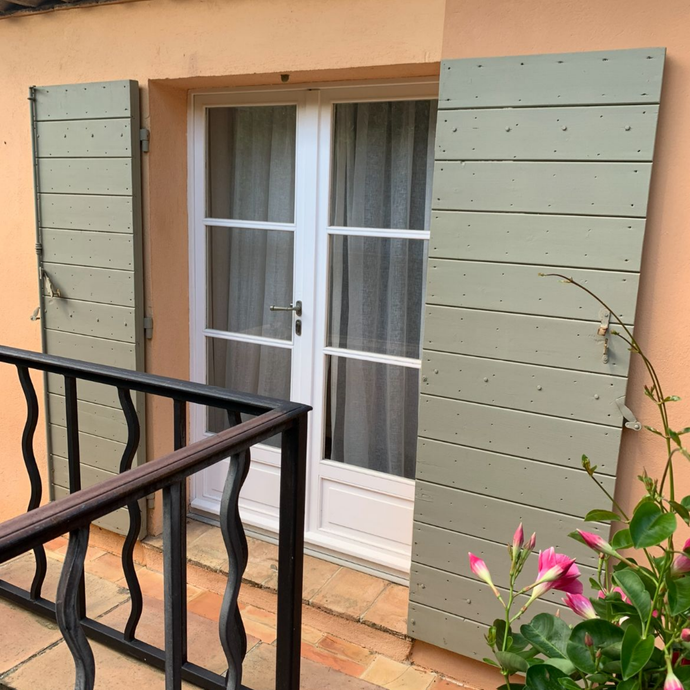 Porte avec volets vert clair, rambarde noire et fleurs roses sur un mur couleur pêche.