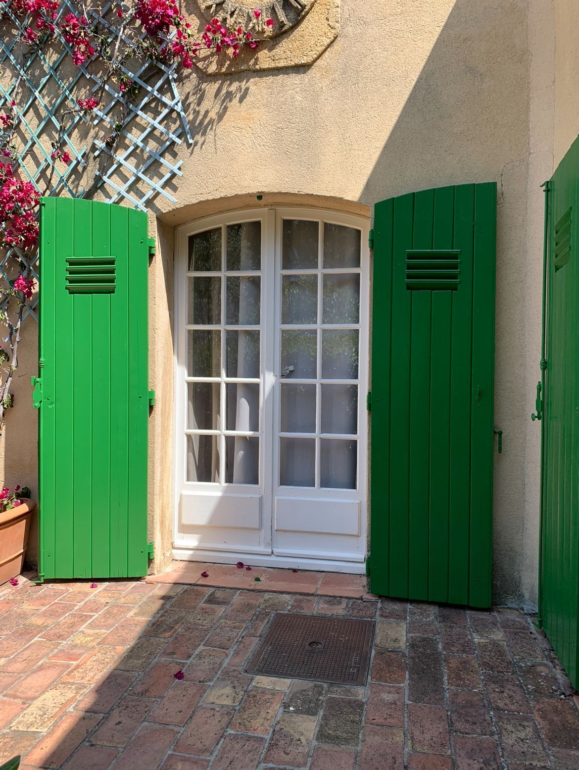Des volets verts encadrent des portes-fenêtres blanches sur un bâtiment beige orné de bougainvilliers.