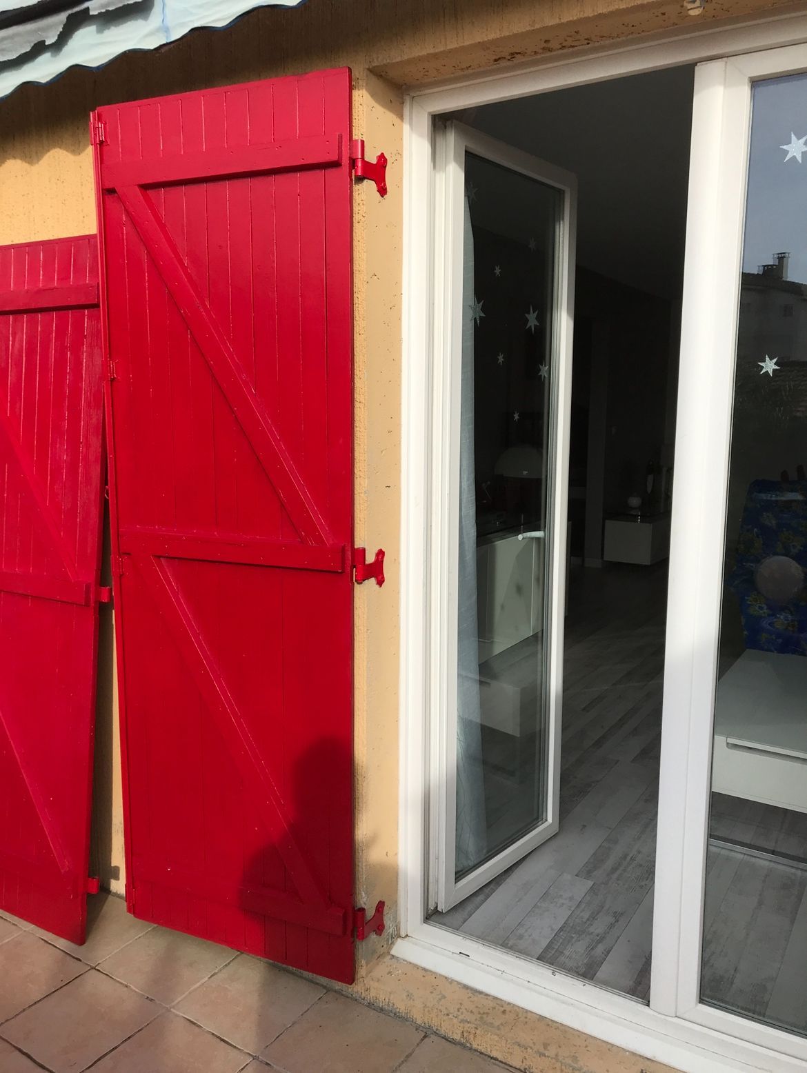 Volets rouges sur un mur jaune, à côté d'une porte vitrée ouverte donnant sur une pièce.