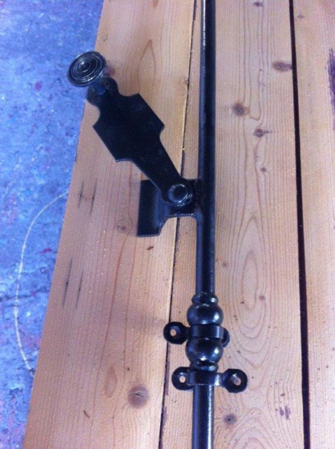 Mât de drapeau en métal noir avec boule décorative et support, posé sur un volet en bois.