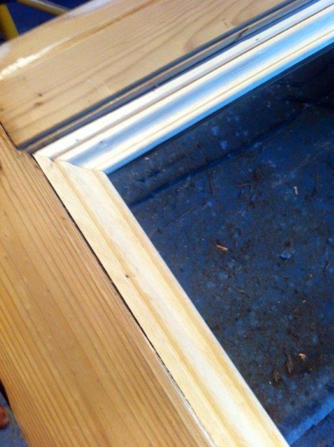 Gros plan sur un cadre de fenêtre en bois naturel avec des finitions blanches. Le cadre est brut et repose sur une surface bleue.