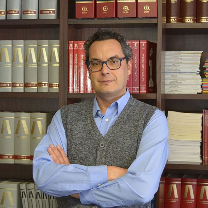 Hombre con gafas, con los brazos cruzados, frente a unas estanterías, vestido con una camisa azul y un suéter gris.