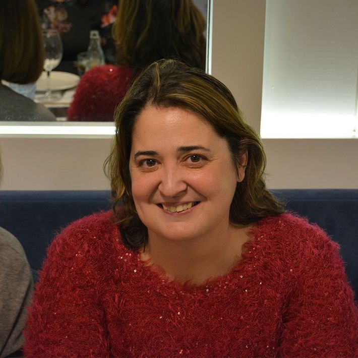Mujer de cabello castaño, sonriente, vistiendo un suéter rojo, sentada en un restaurante.