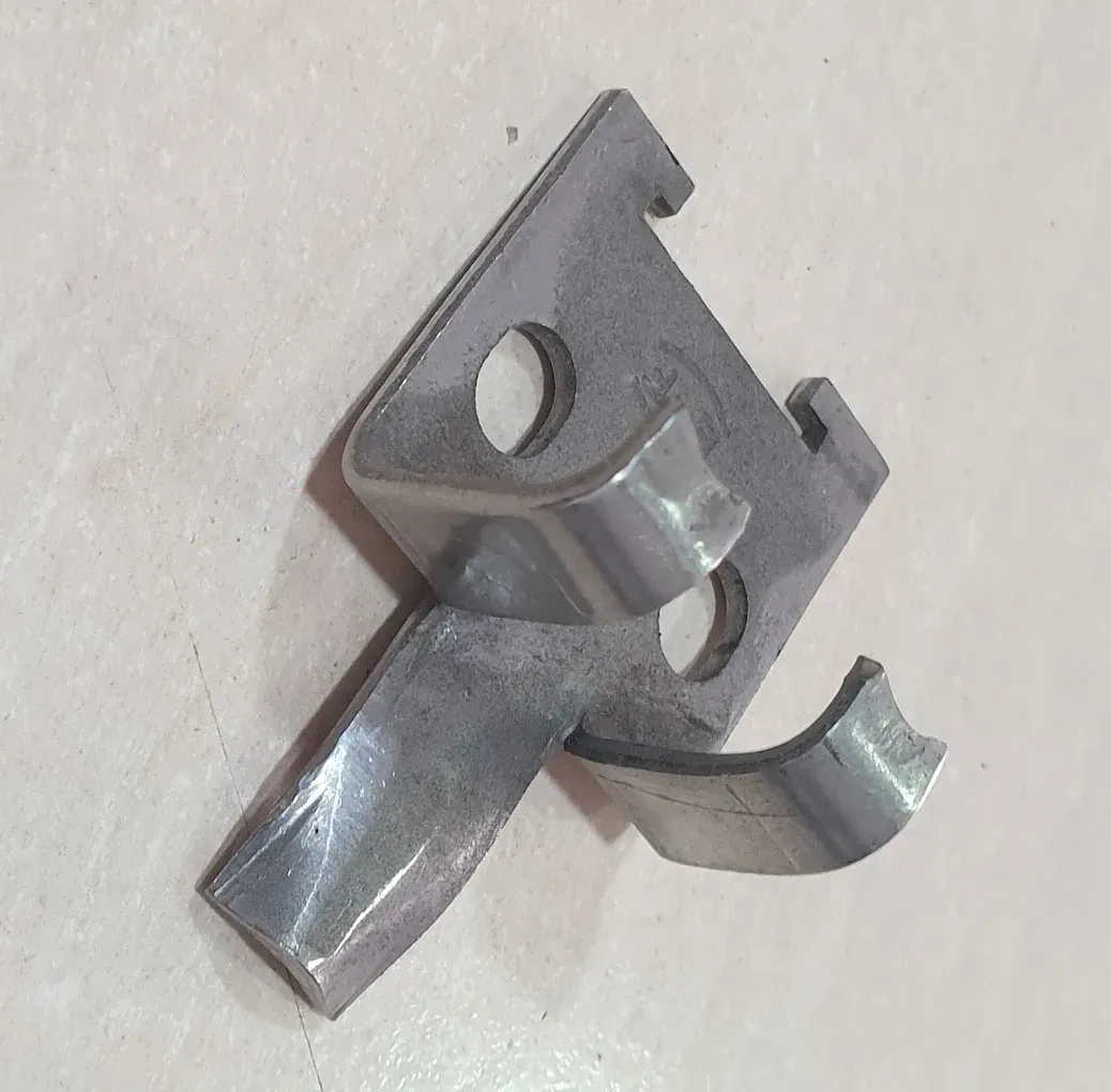 Clip metálico con bordes doblados y dos agujeros, apoyado sobre una superficie blanca.