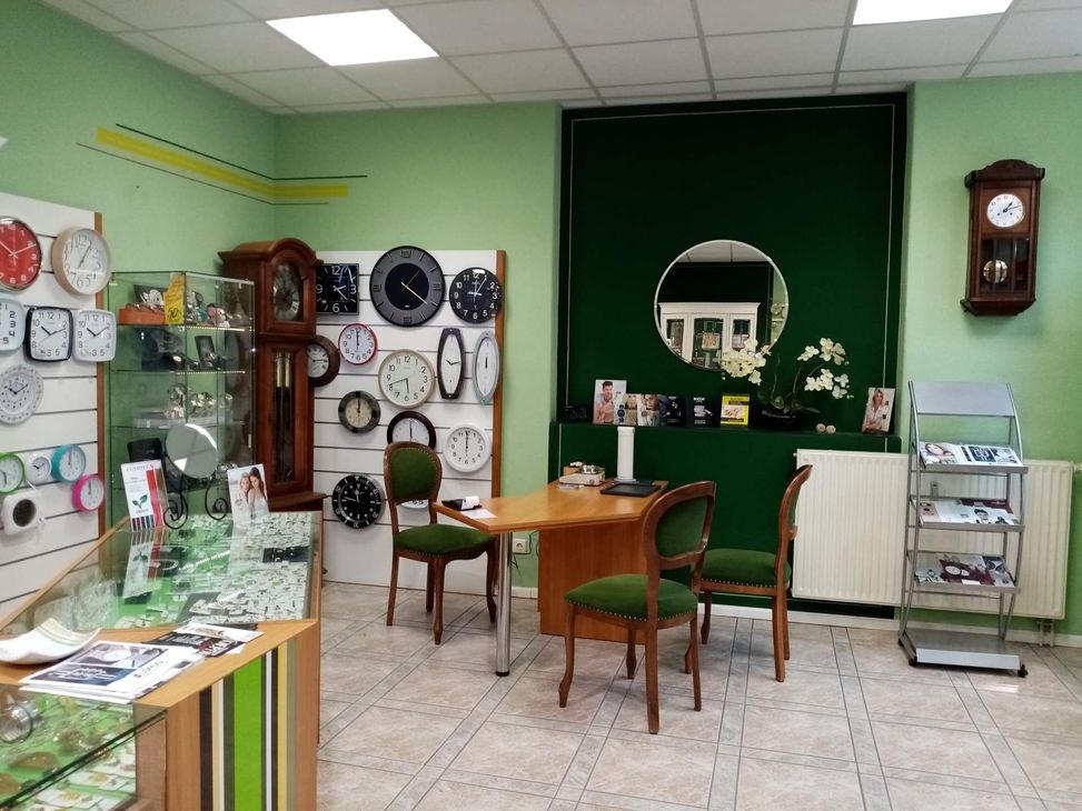 Verschiedenen Uhrendesigns an der Wand, grünen Akzenten, einem Holztisch und Stühlen in einem gemütlichen Ausstellungsbereich.